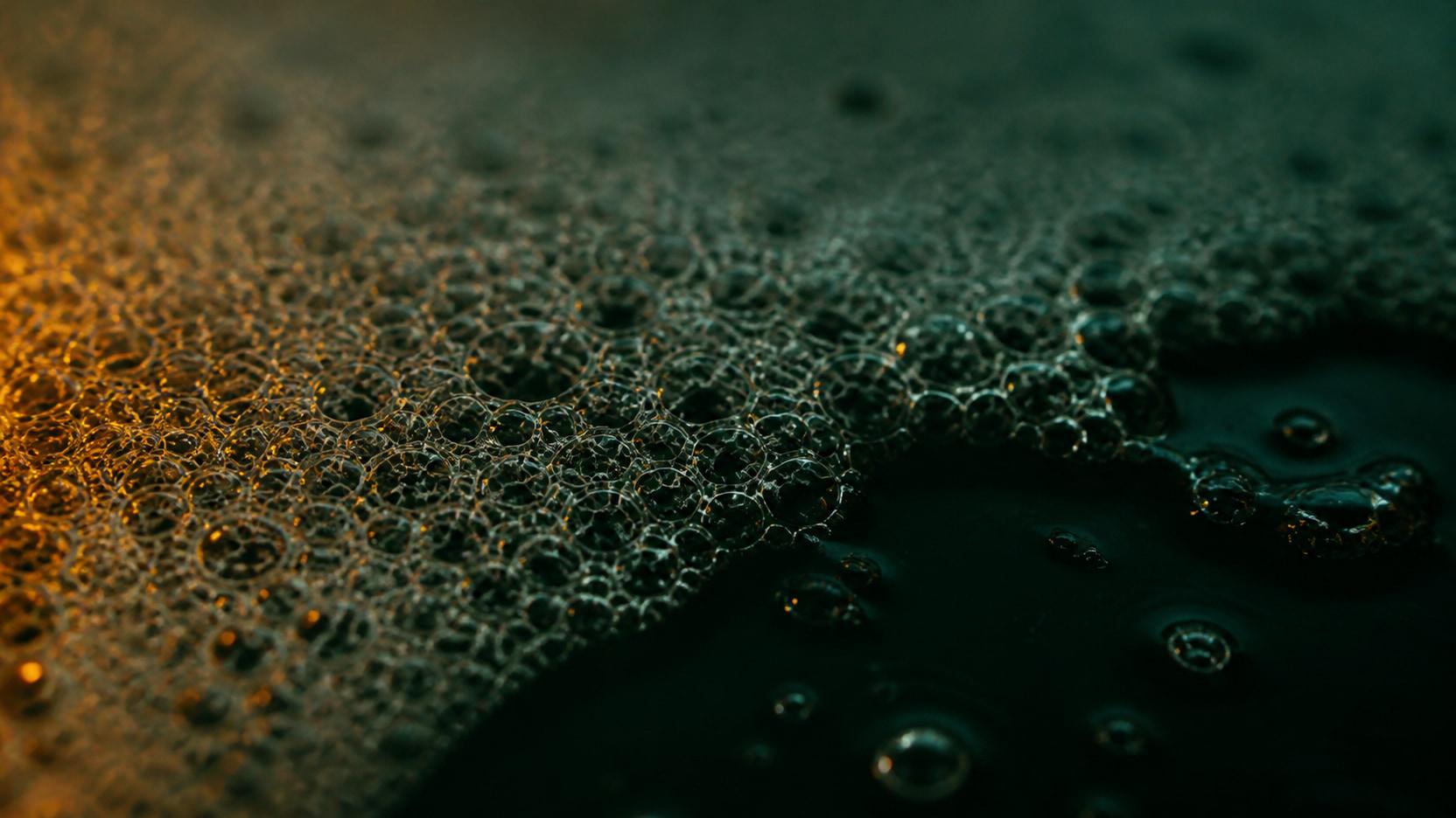 Macro cross-section of dense manure foam, thousands of tight bubbles collapsing at one edge to reveal the dark liquid below.